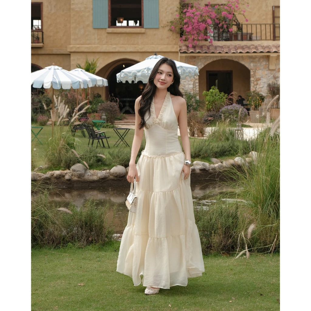 Woman in a long white dress standing in a garden with a building and umbrellas in the background.
