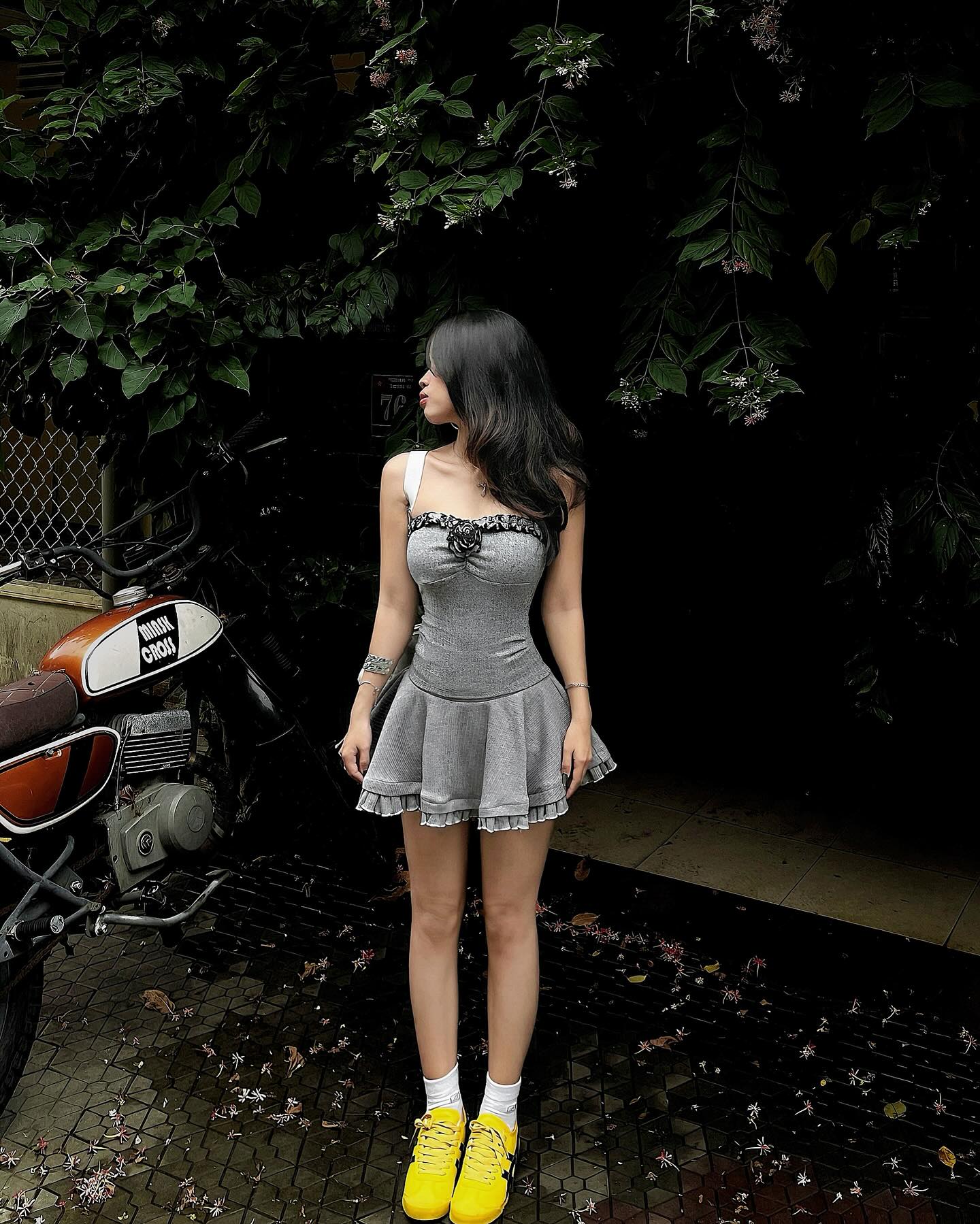 Woman in a gray dress standing next to a motorcycle in a dark, outdoor setting