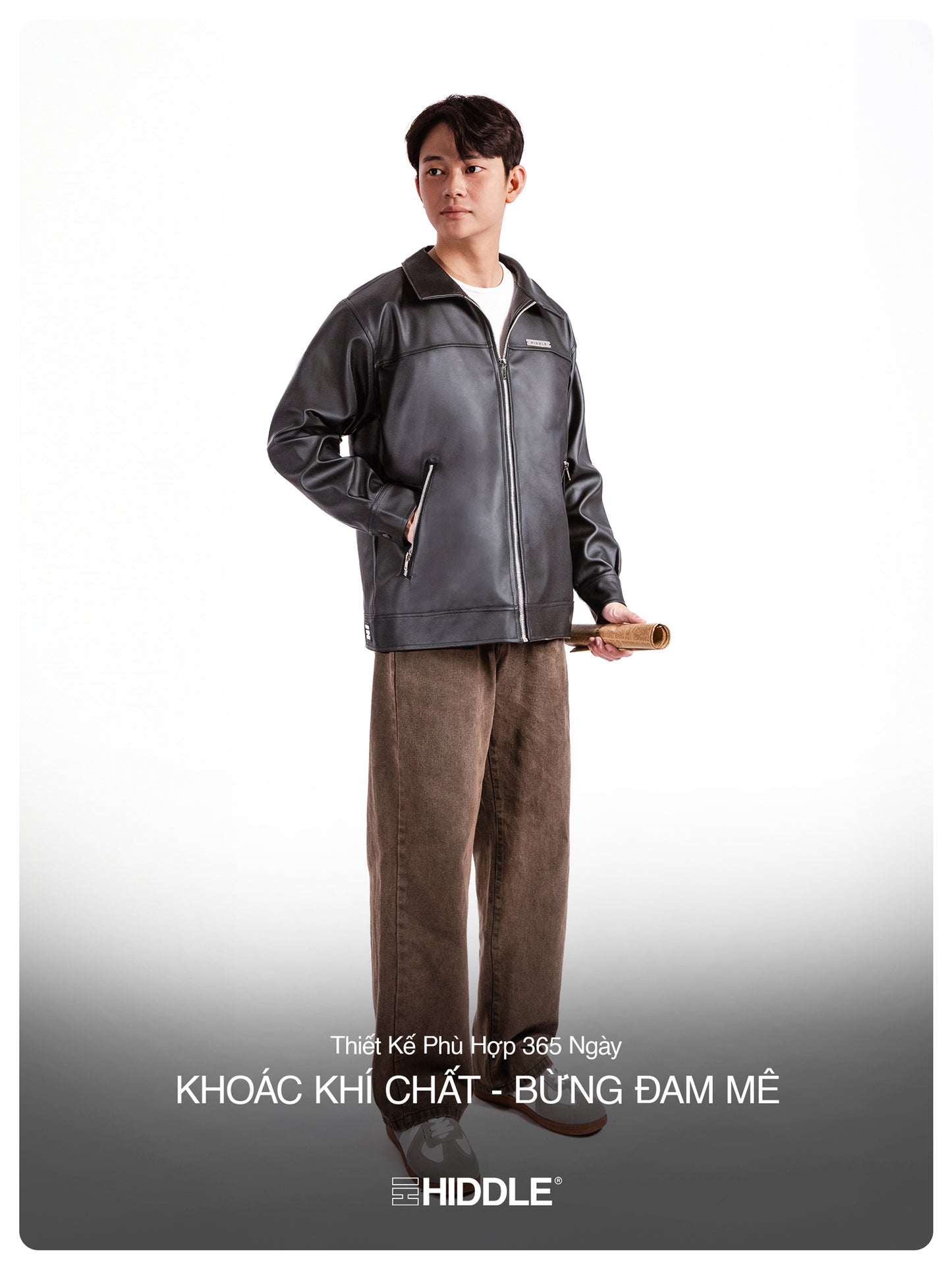 Person wearing a gray jacket and brown pants holding a rolled-up paper on a white background with Hiddle branding.
