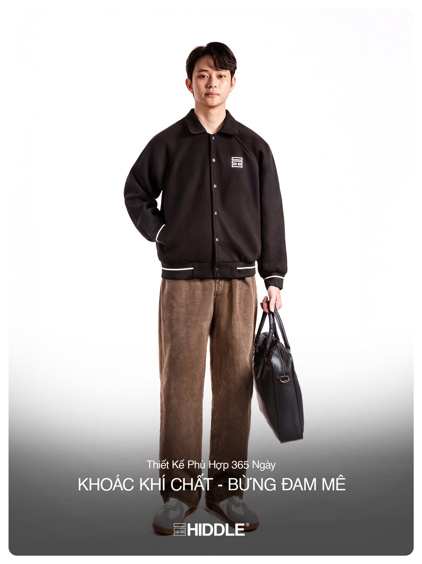 Man wearing a black varsity jacket and brown pants holding a black bag on a white background