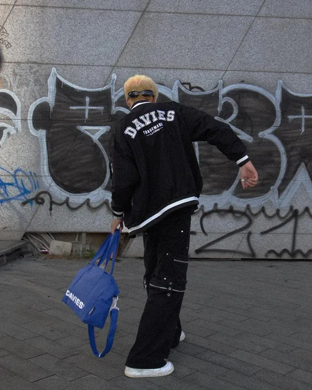 Person wearing a black 'DAVIES' jacket with a blue bag, standing in front of a graffiti-covered wall.