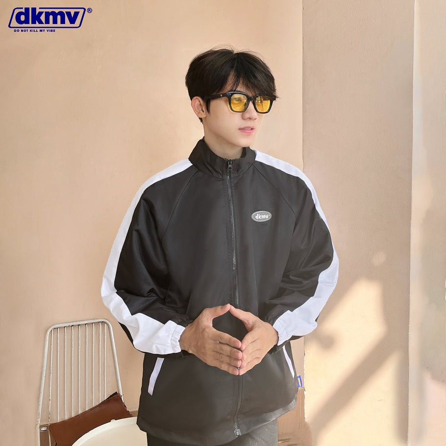 Person wearing a black and white jacket with a visible brand logo, sitting against a beige wall.