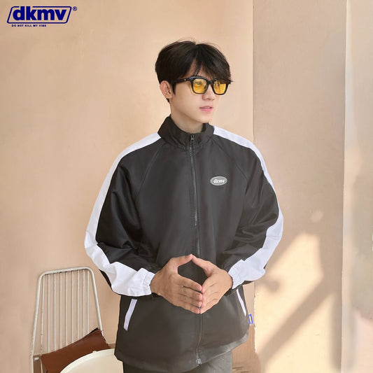 Person wearing a black and white jacket with a visible brand logo, sitting against a beige wall.