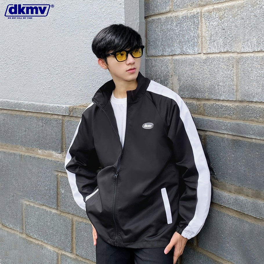 Person wearing a black and white jacket leaning against a gray brick wall with DKMV logo.