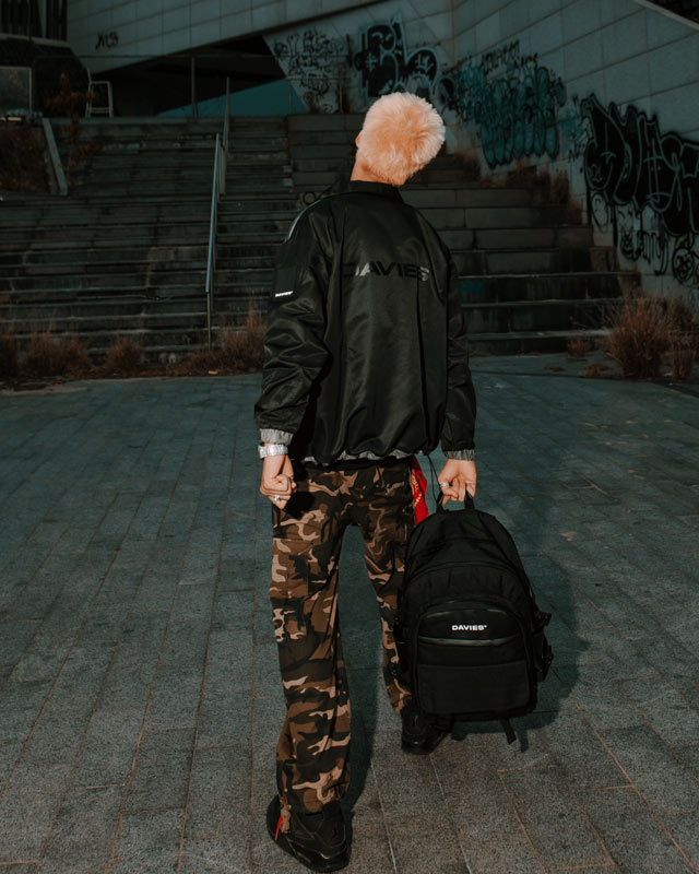 Person wearing a black jacket and camouflage pants holding a black backpack in an urban setting with graffiti.