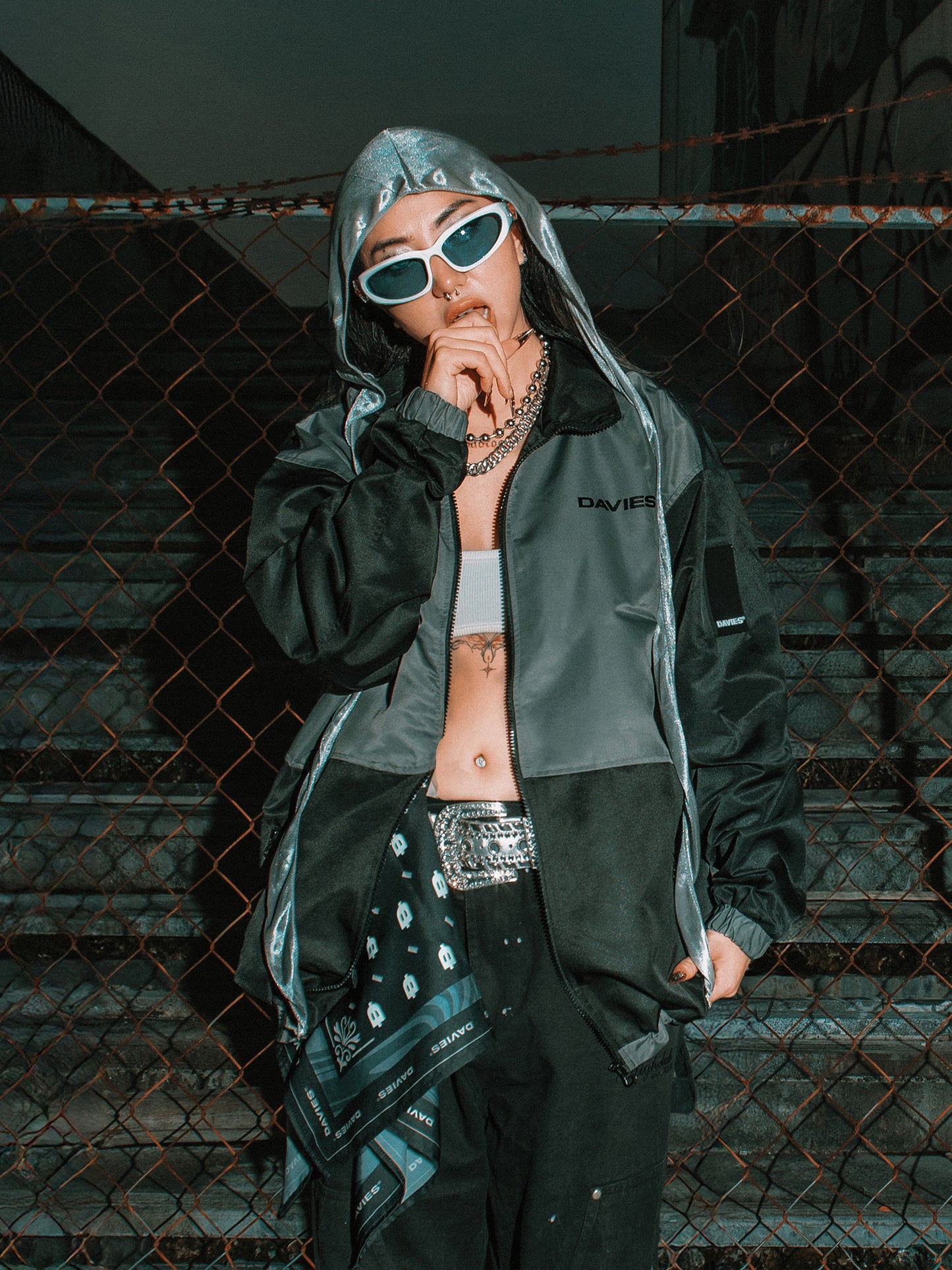 Person wearing a green raincoat with a hood, sunglasses, and a chain necklace in front of a chain-link fence.