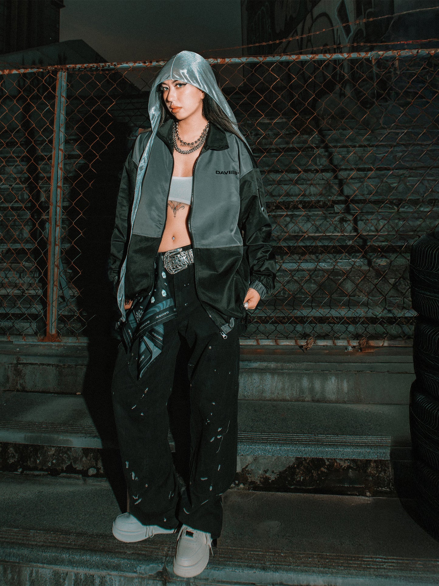 Person wearing a dark jacket and pants standing against a chain-link fence.