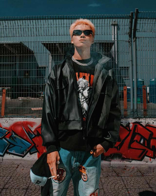 Person with pink hair wearing sunglasses, black jacket, and jeans in front of a graffiti wall.