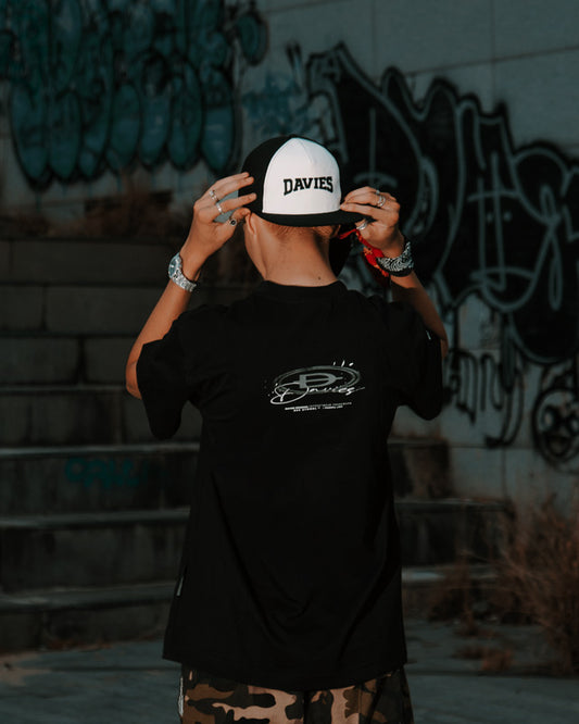 Person wearing a black t-shirt with a logo and a white cap with 'DAVIES' branding, standing against a graffiti-covered wall.
