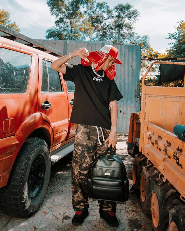Person holding a black backpack in front of orange trucks with a red bandana on their head.
