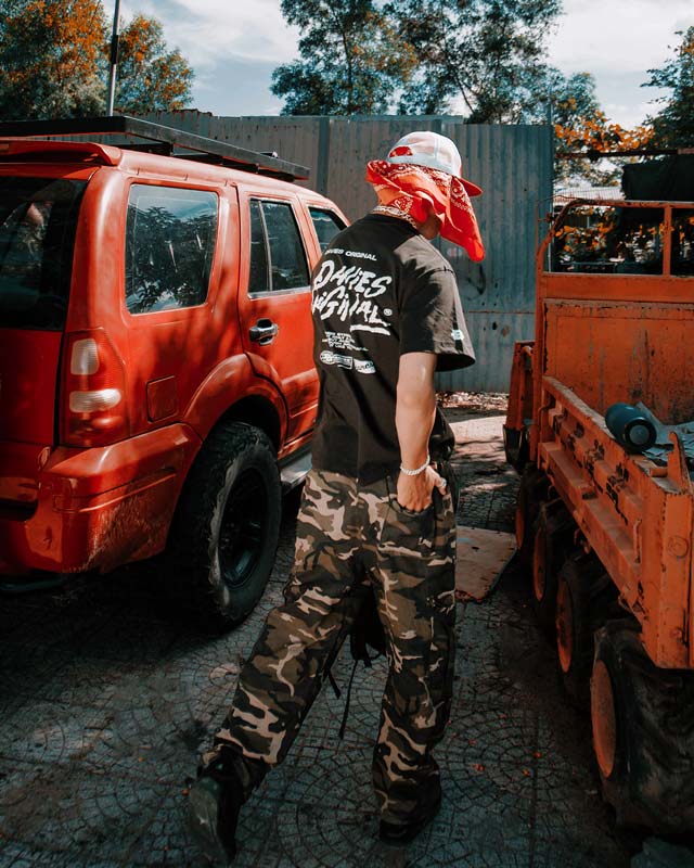 Person wearing a black t-shirt with white text and camouflage pants standing near a red SUV and orange truck.