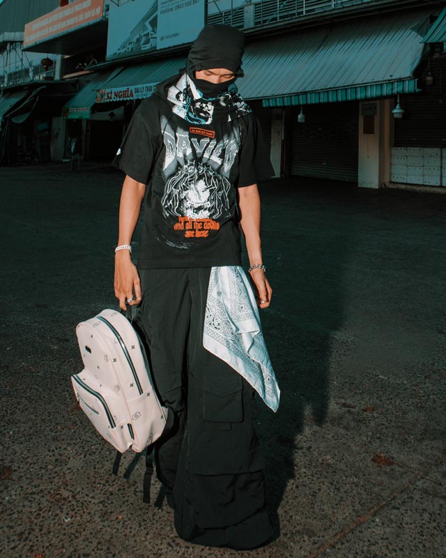 Person wearing a black outfit with a graphic t-shirt, holding a white backpack in an urban setting.