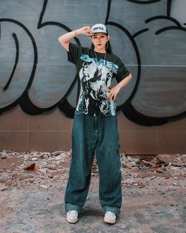 Person wearing a graphic t-shirt and jeans standing in front of a graffiti-covered wall.