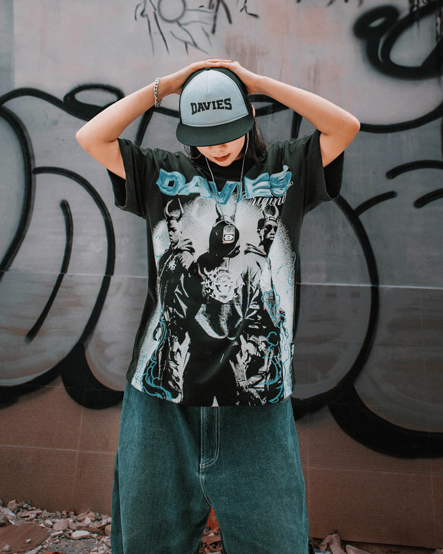 Person wearing a graphic t-shirt and cap with 'DAVIES' brand, standing against a graffiti-covered wall.