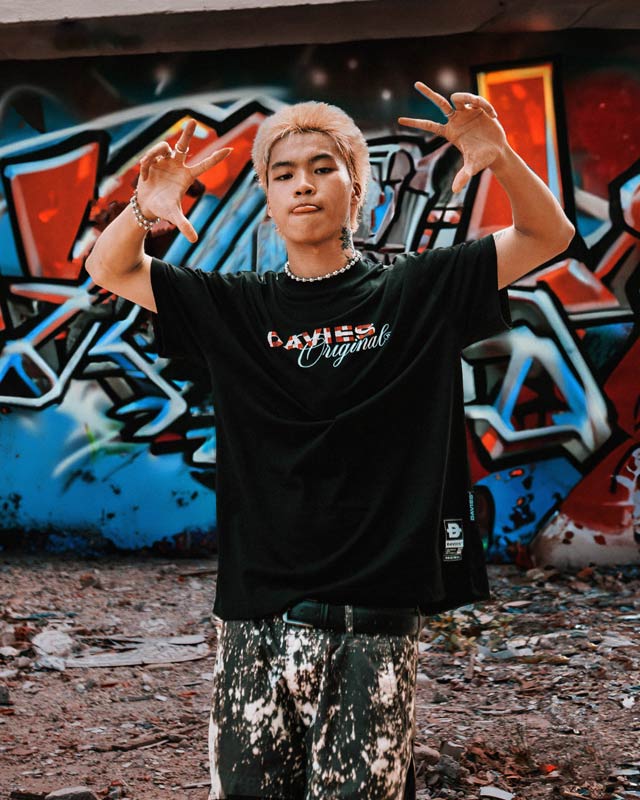 Person wearing a black t-shirt with visible branding in front of a graffiti-covered wall.