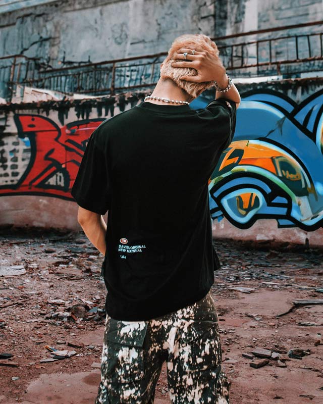 Person wearing a black t-shirt with a logo, standing in front of graffiti art.