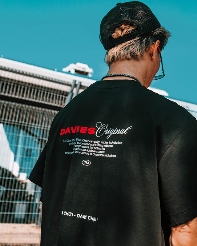 Person wearing a black t-shirt with 'DAVIES Original' branding, standing against a blue sky.