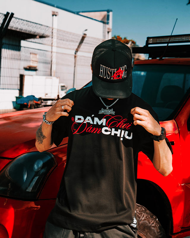 Person wearing a black cap and t-shirt with text, standing in front of a red car.