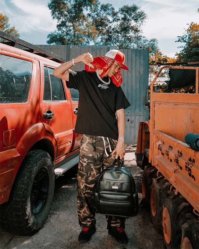 Person holding a backpack in front of orange vehicles with a blue wall and trees in the background