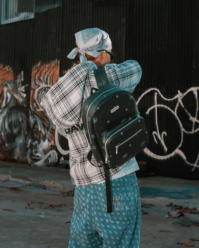 Person wearing a plaid shirt, blue pants, and a black backpack with a bandana in an urban setting with graffiti.