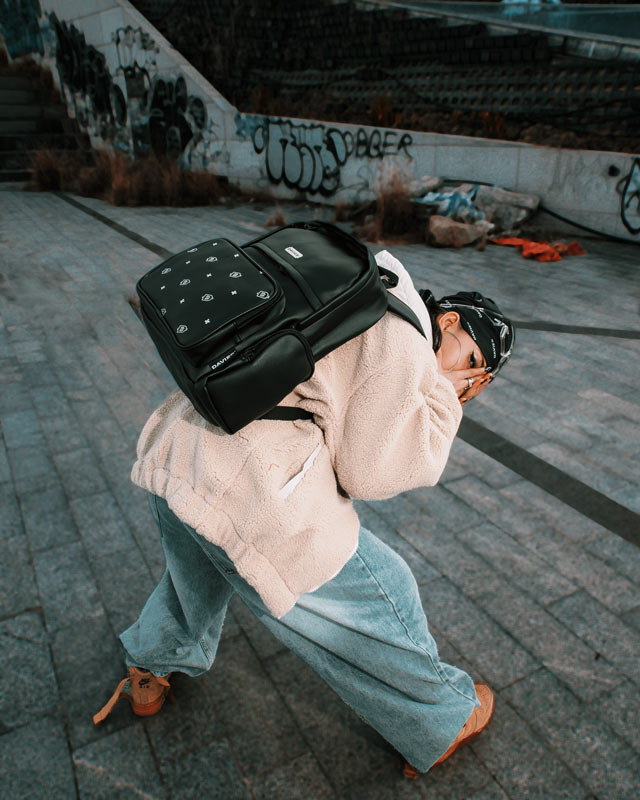Person wearing a black backpack with a unique design, standing on a concrete surface with graffiti in the background.