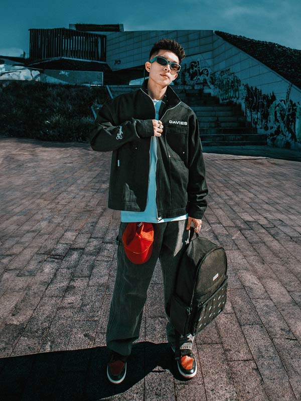 Person wearing a black jacket with a brand logo, holding a red bag and black suitcase, standing on a brick pavement.