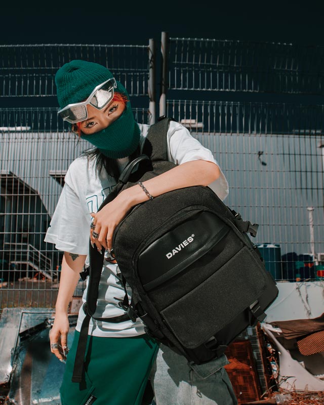 Person holding a black backpack with 'DAVIES' branding against a metal fence background