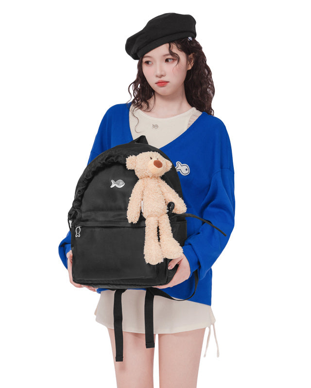 Person holding a black backpack with a teddy bear attached, wearing a blue sweater and black beret.