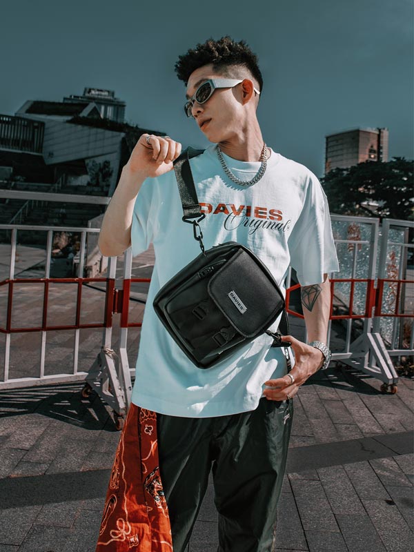 Person wearing a light blue t-shirt with 'DAVIES' branding, holding a black bag in an urban setting.