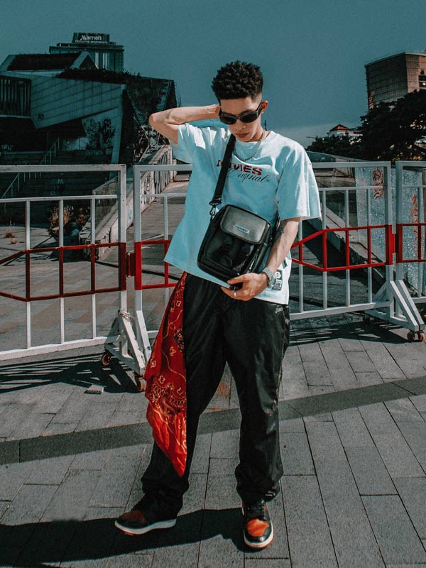 Person wearing a light blue t-shirt with a graphic design, dark pants, and sunglasses, standing on a rooftop with buildings in the background.