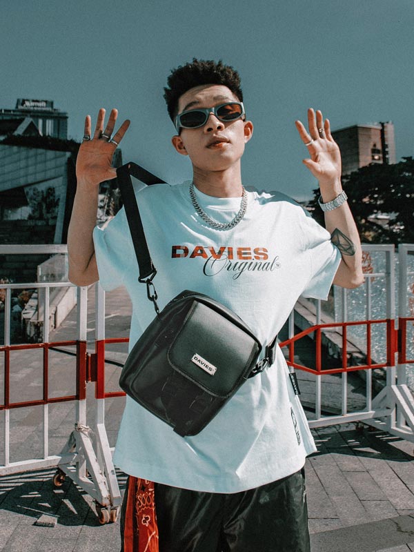 Person wearing a light blue t-shirt with 'DAVIES Original' text, carrying a black bag outdoors.