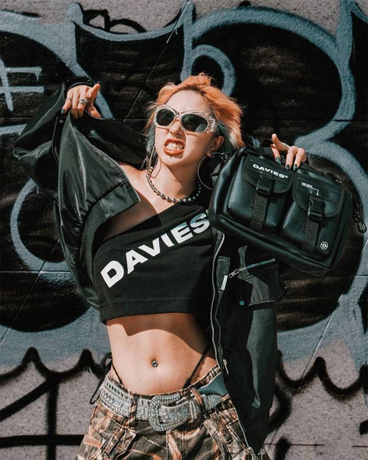 Person wearing a black 'DAVIES' shirt and holding a black bag against a graffiti-covered wall.
