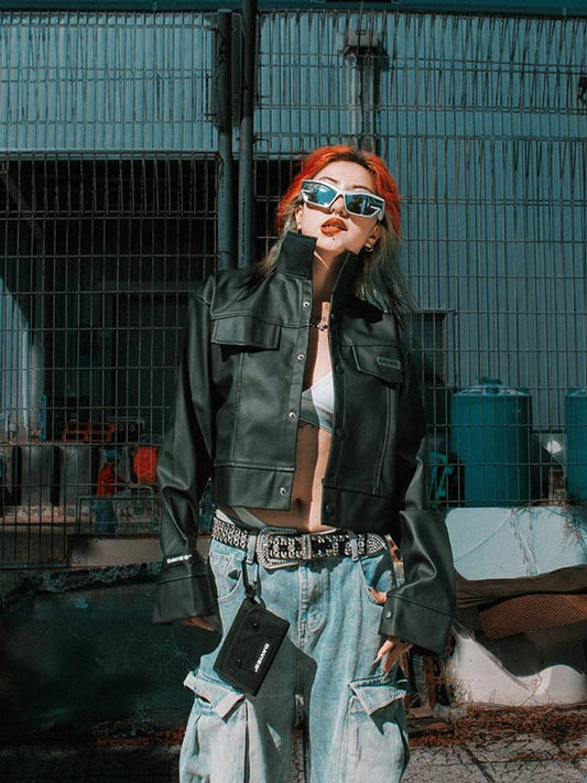 Person wearing a green leather jacket and blue jeans with sunglasses, standing in an industrial setting.