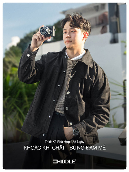 Man taking a photo with a camera, wearing a dark jacket and sweater, with a blurred outdoor background.