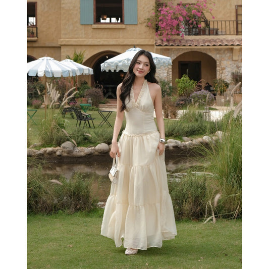 Woman in a long white dress standing in a garden with a building and umbrellas in the background.