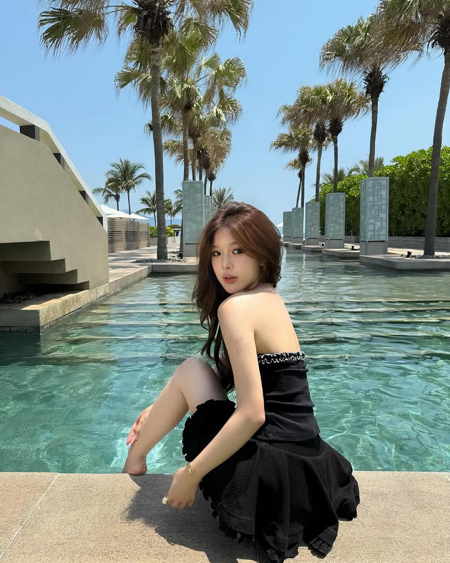 Woman in a black dress sitting by a pool with palm trees in the background