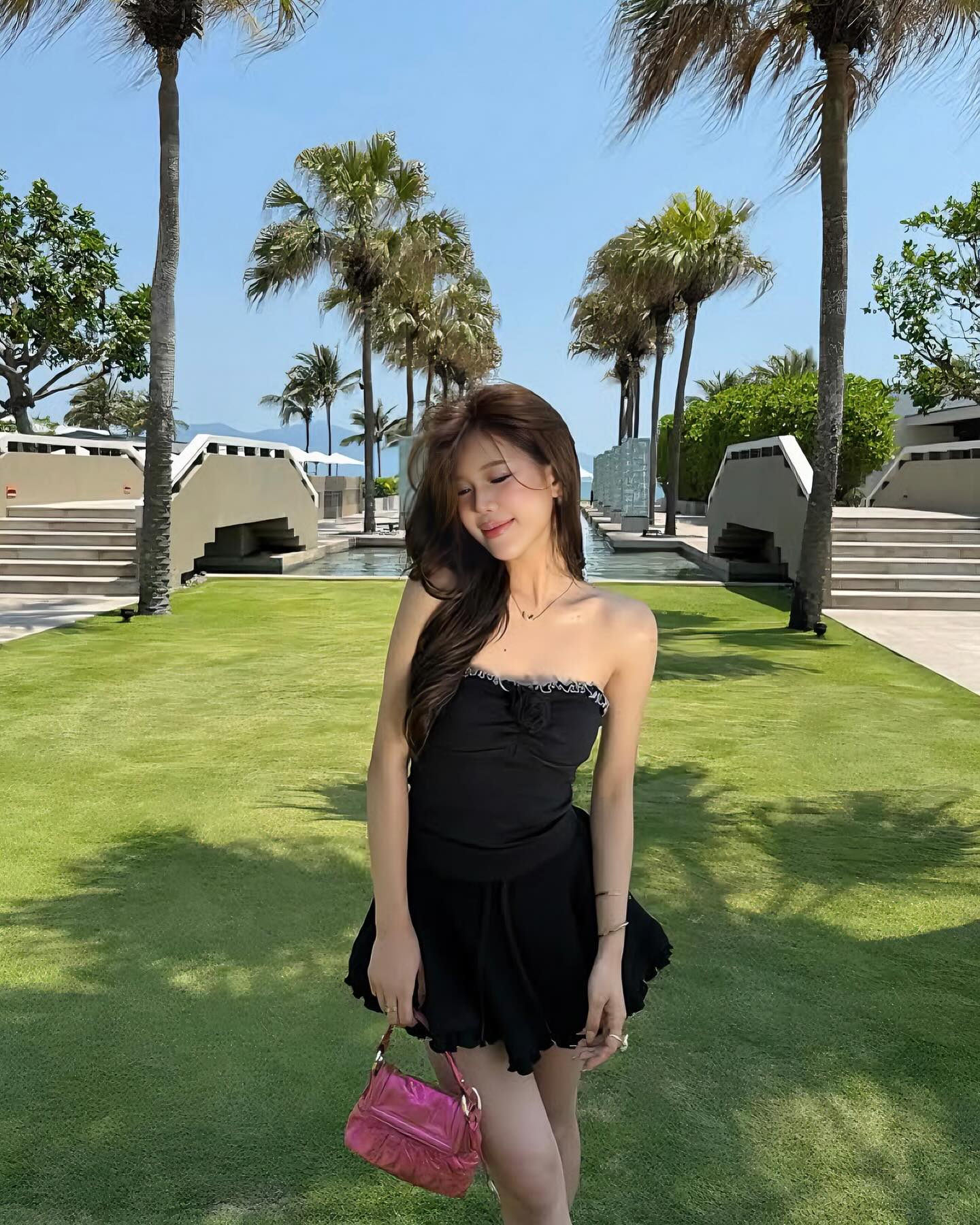Woman in a black dress holding a pink bag on a grassy area with palm trees in the background