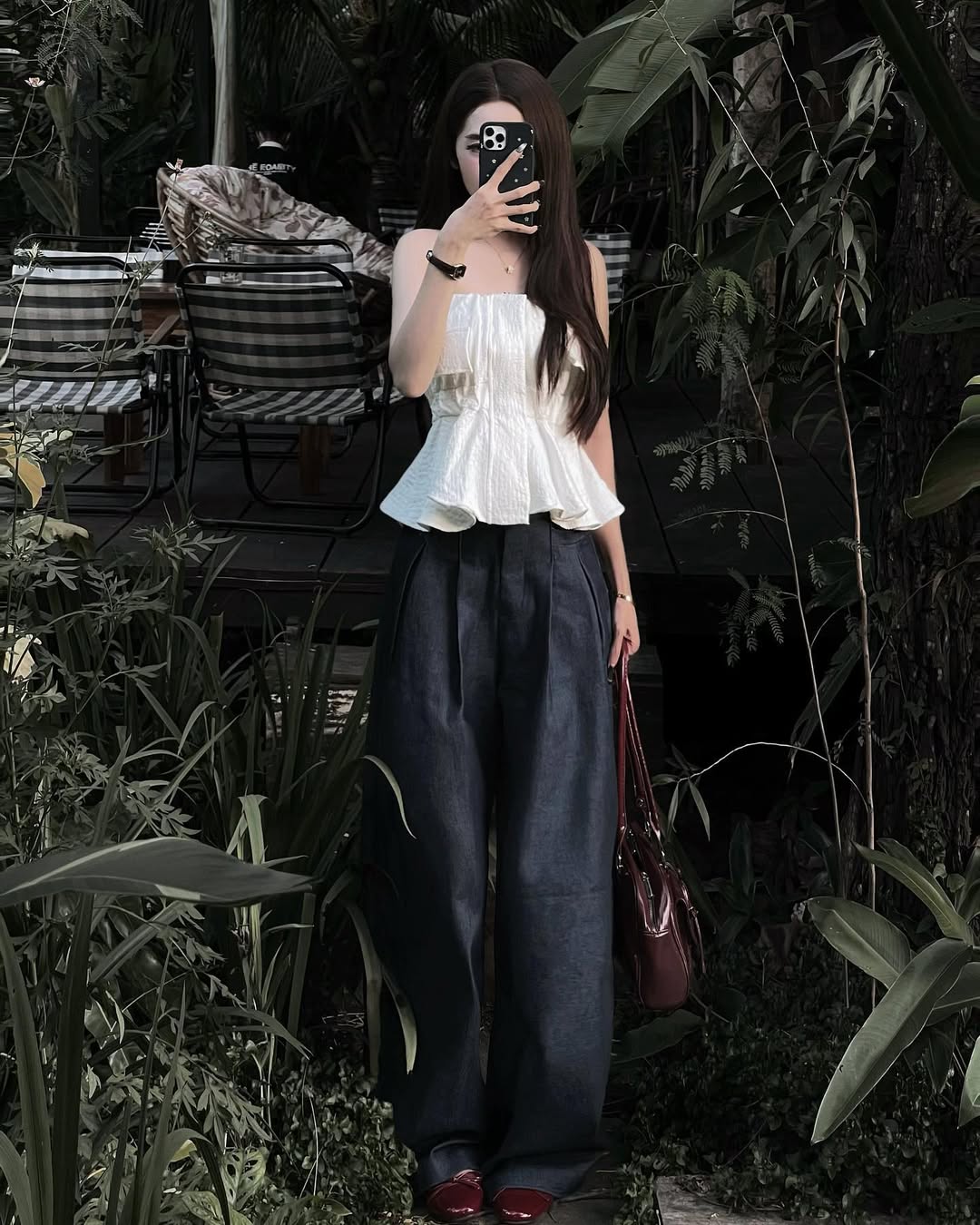 Woman taking a selfie in a white top and dark pants amidst greenery