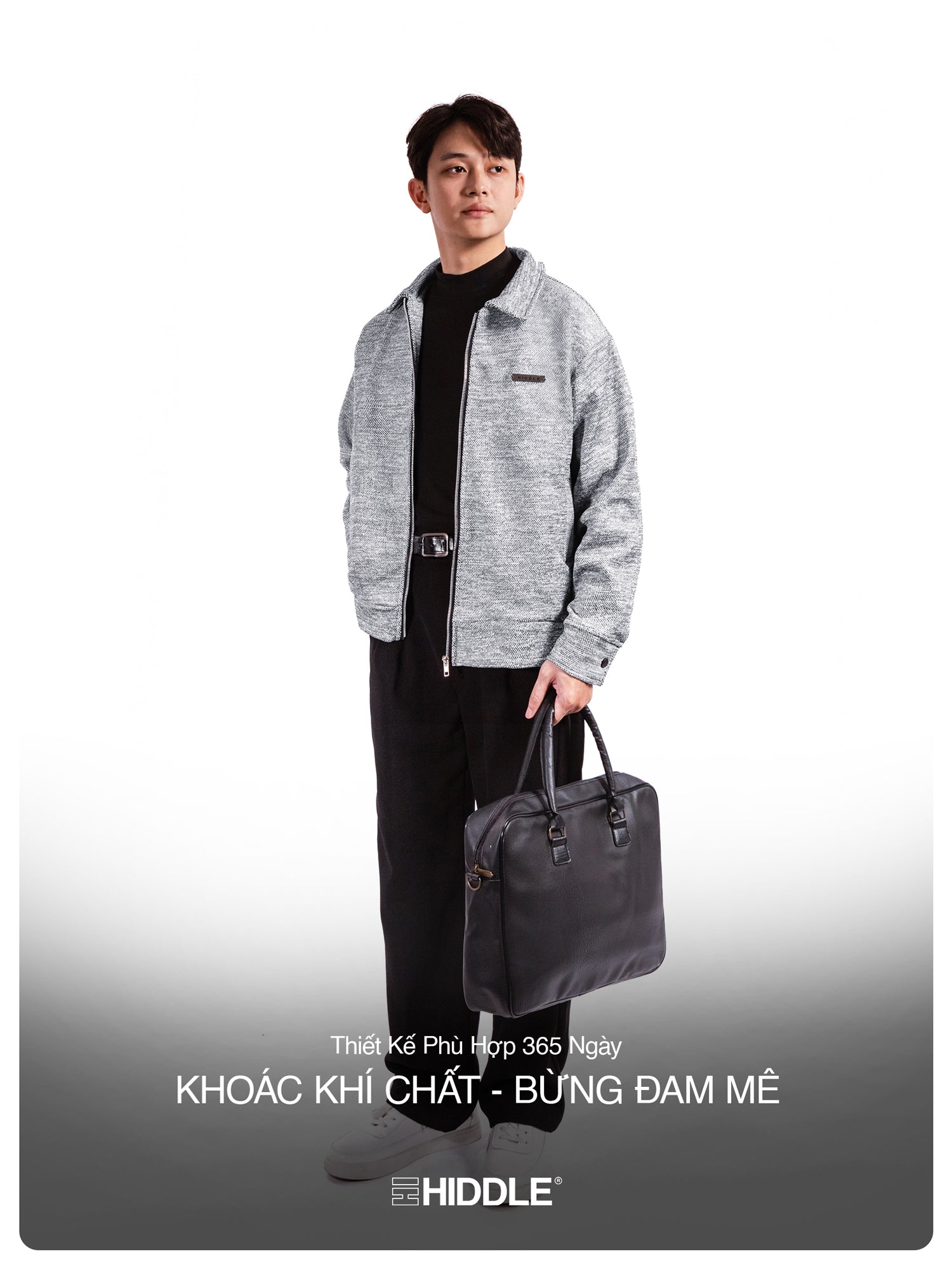 Man holding a black bag wearing a gray jacket and black pants on a white background with Hiddle branding.