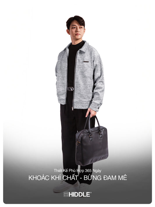 Man holding a black bag wearing a gray jacket and black pants on a white background with Hiddle branding.