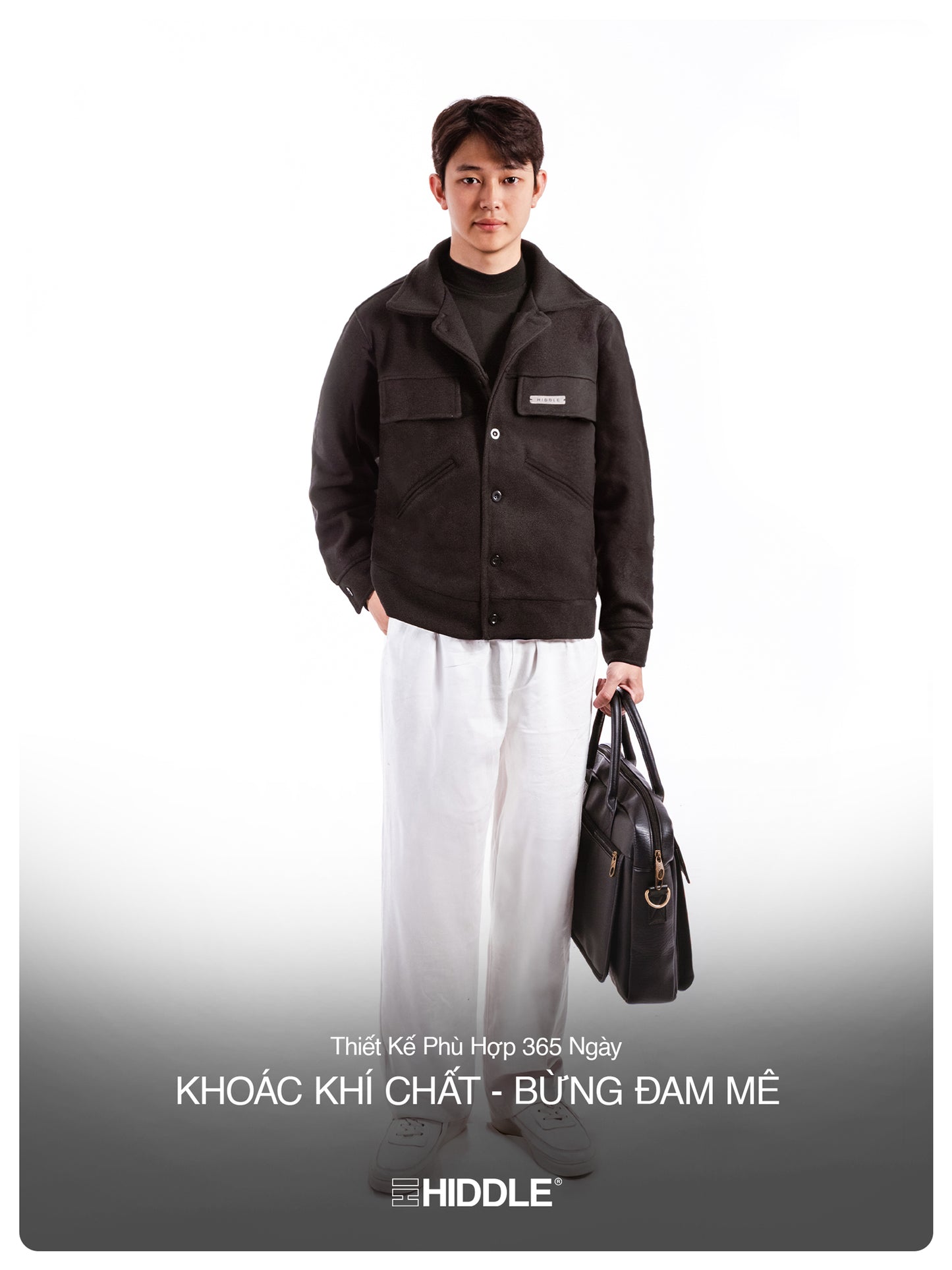 Man wearing a dark jacket and white pants holding a black bag on a white background
