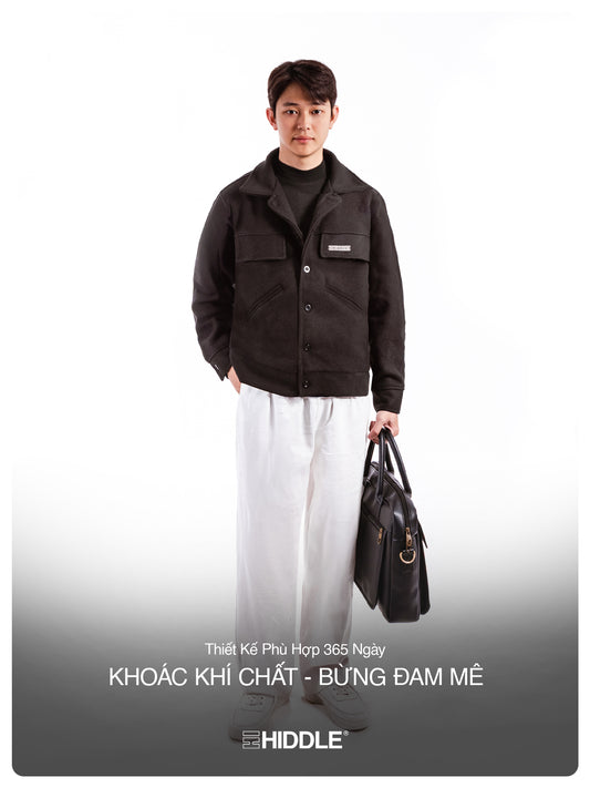 Man wearing a dark jacket and white pants holding a black bag on a white background