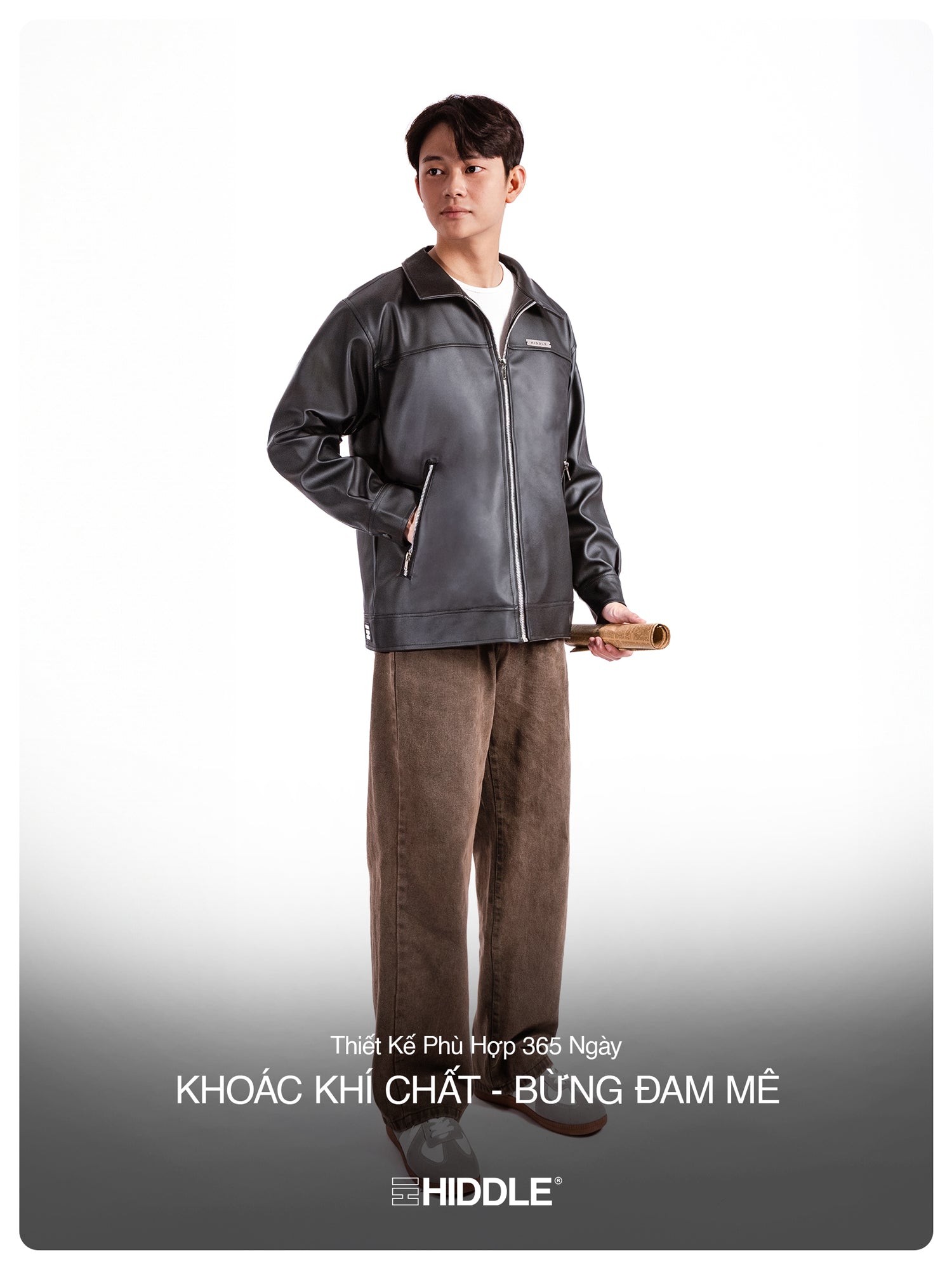 Person wearing a gray jacket and brown pants holding a rolled-up paper on a white background with Hiddle branding.