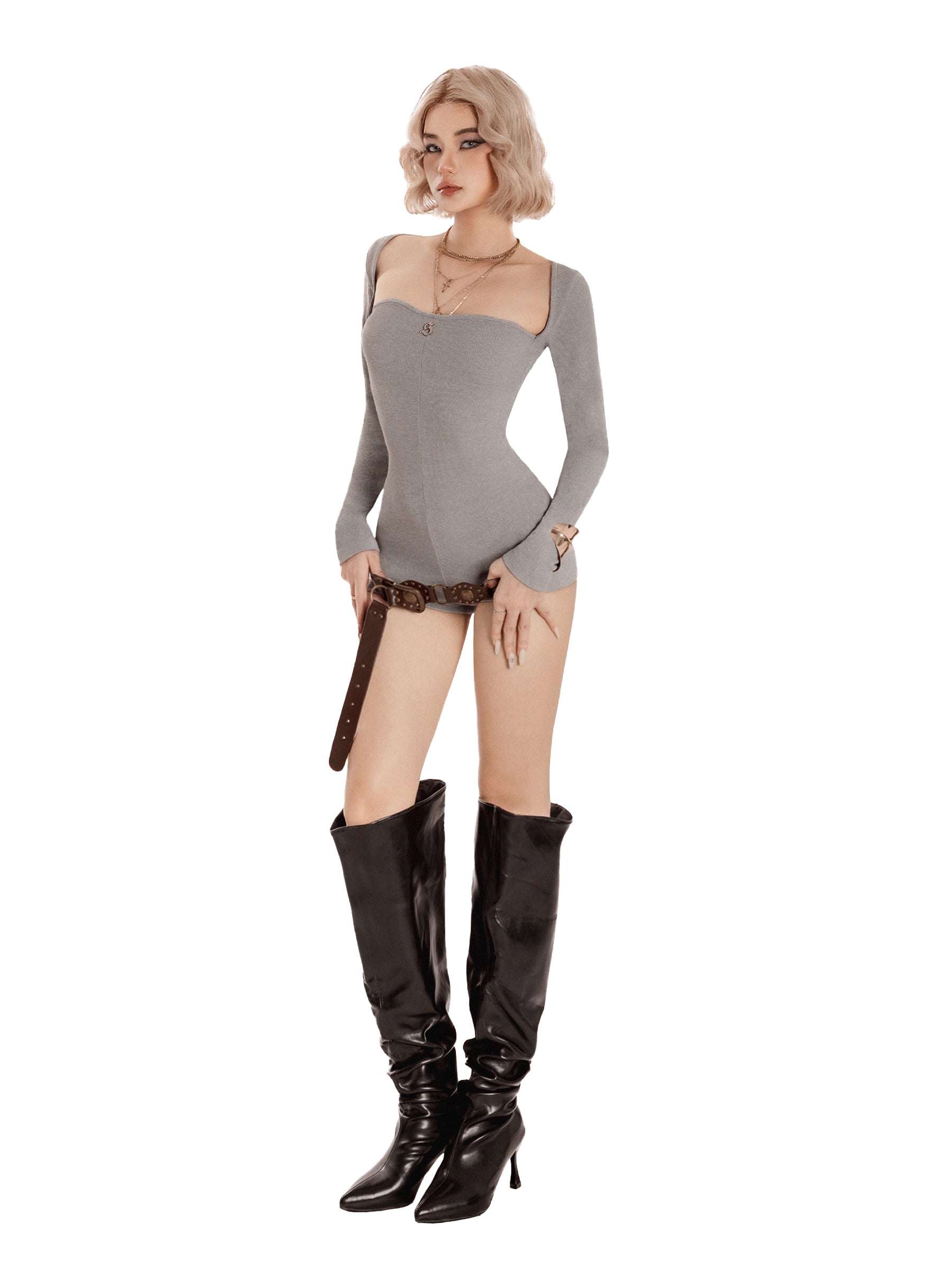 Woman wearing a gray bodysuit and black knee-high boots holding a whip on a white background