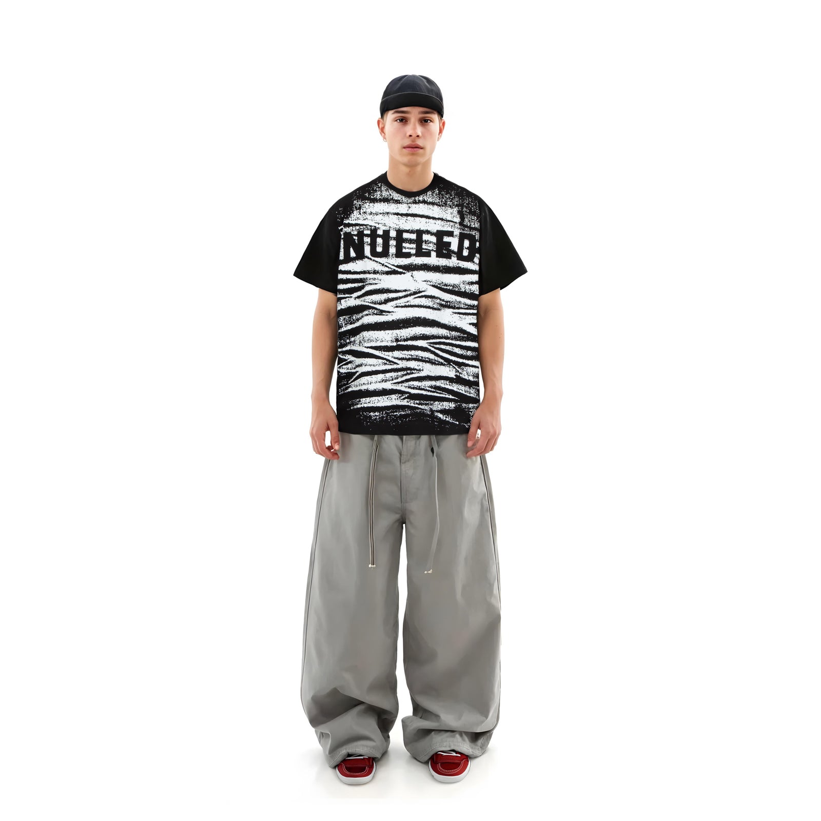 Person wearing a black and white graphic t-shirt with text, gray pants, and red shoes on a white background
