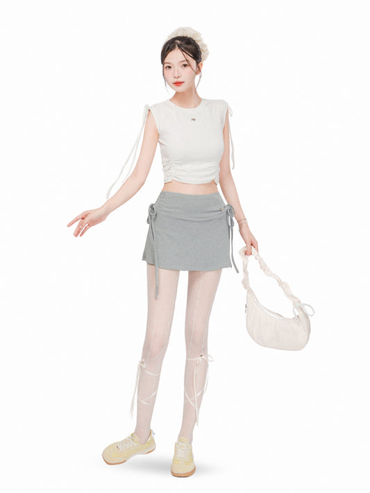 Woman wearing a white sleeveless top and gray skirt with white tights and shoes, holding a beige handbag on a white background.
