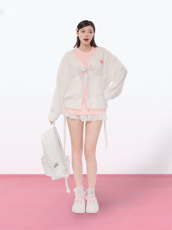 Woman wearing a light gray cardigan over a pink top with white shorts, holding a white backpack on a pink floor and white background.