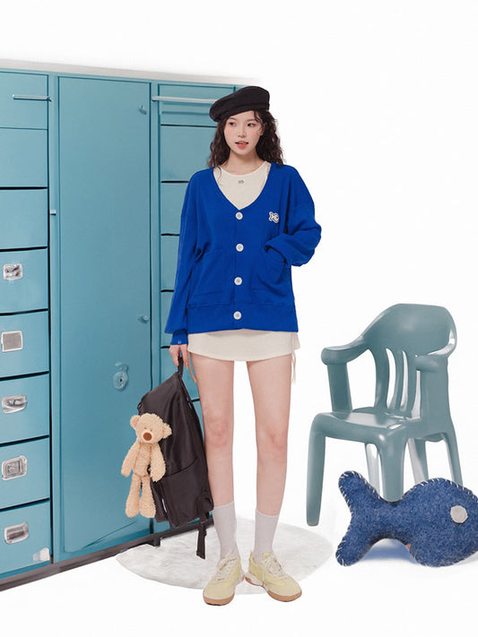 Person wearing a blue cardigan in front of blue lockers with a teddy bear and chair in the foreground.