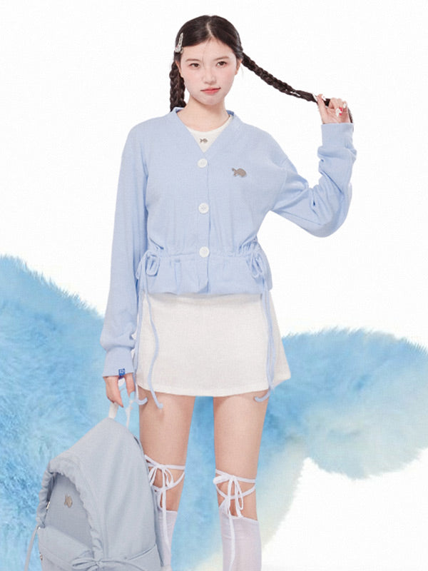 Woman wearing a light blue cardigan over a white top and skirt, with a fluffy gray bag on a white background.