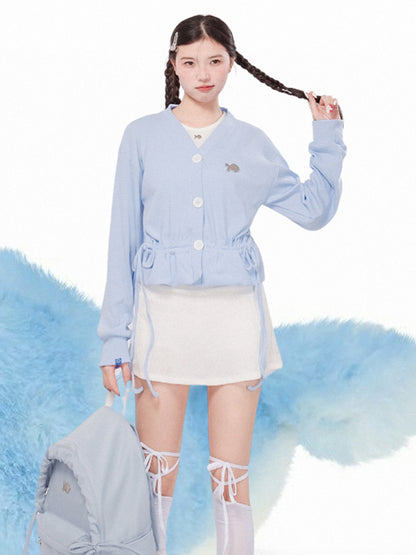 Woman wearing a light blue cardigan over a white top and skirt, with a fluffy gray bag on a white background.